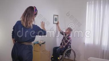 轮椅上的残疾丈夫选择在白色墙上的相框图片的位置，而她可爱的妻子则塑造了